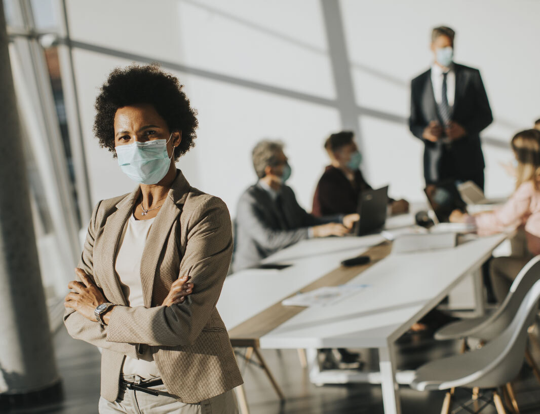 Woman in business attire with her arms crossed wearing a mask at the end of a boardroom table. Another group of people gather around the end of the table.