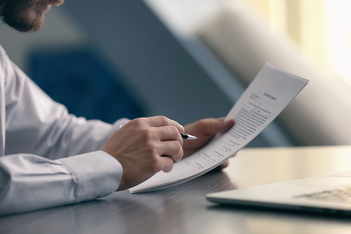 Man wearing a dress shirt and sitting at a table is holding a pen to point at the piece of paper he's reading.